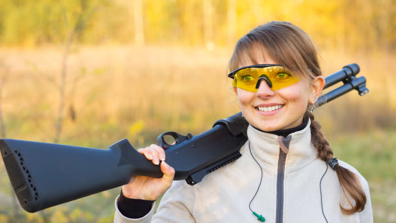 Young girl smiles while carrying a shotgun in a field over her shoulder.